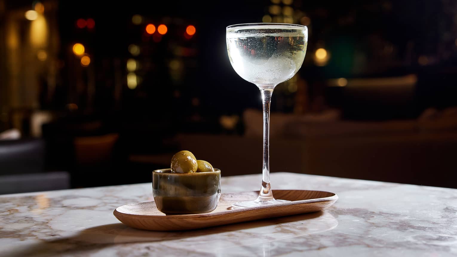 Clear cocktail in tall wine glass and bowl of green olives in wooden tray on marble table