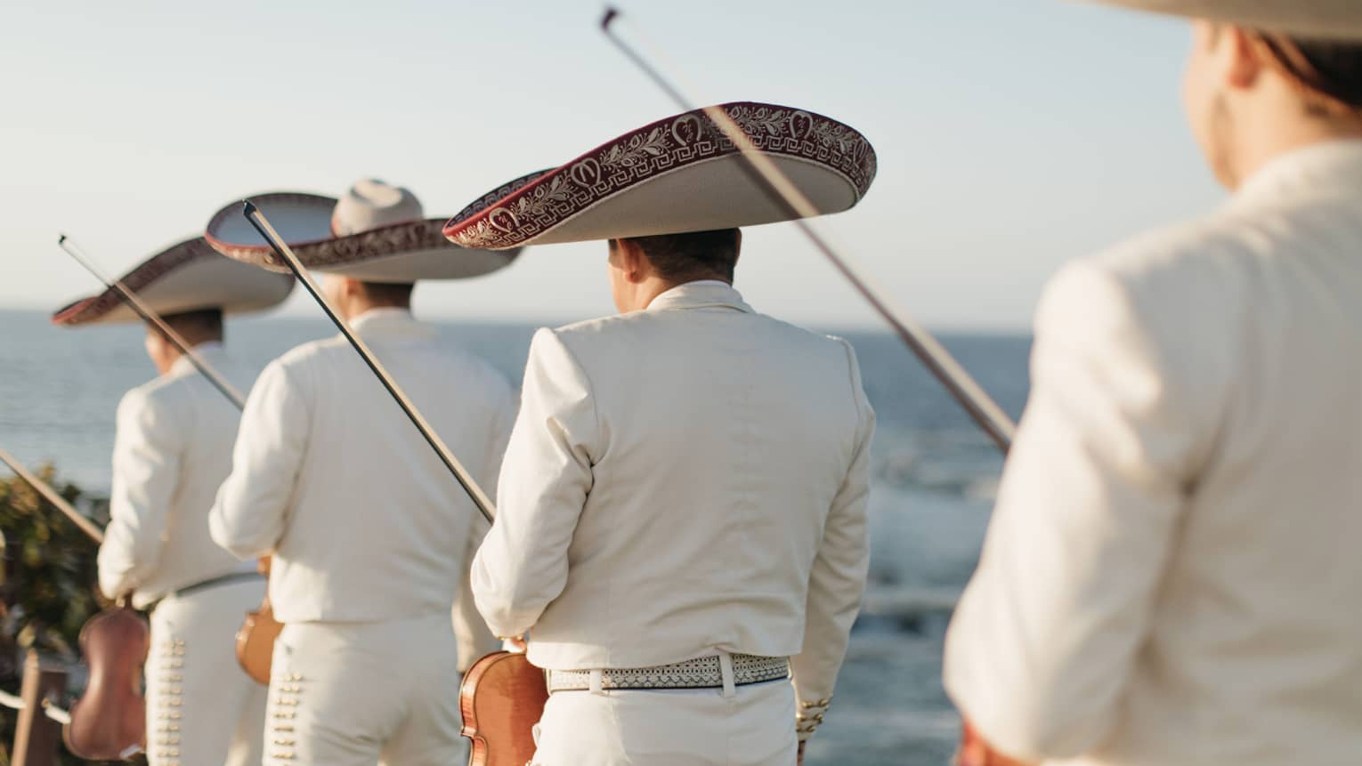 The backs of four men in white suits and large hats holding violins with the ocean in the background.
