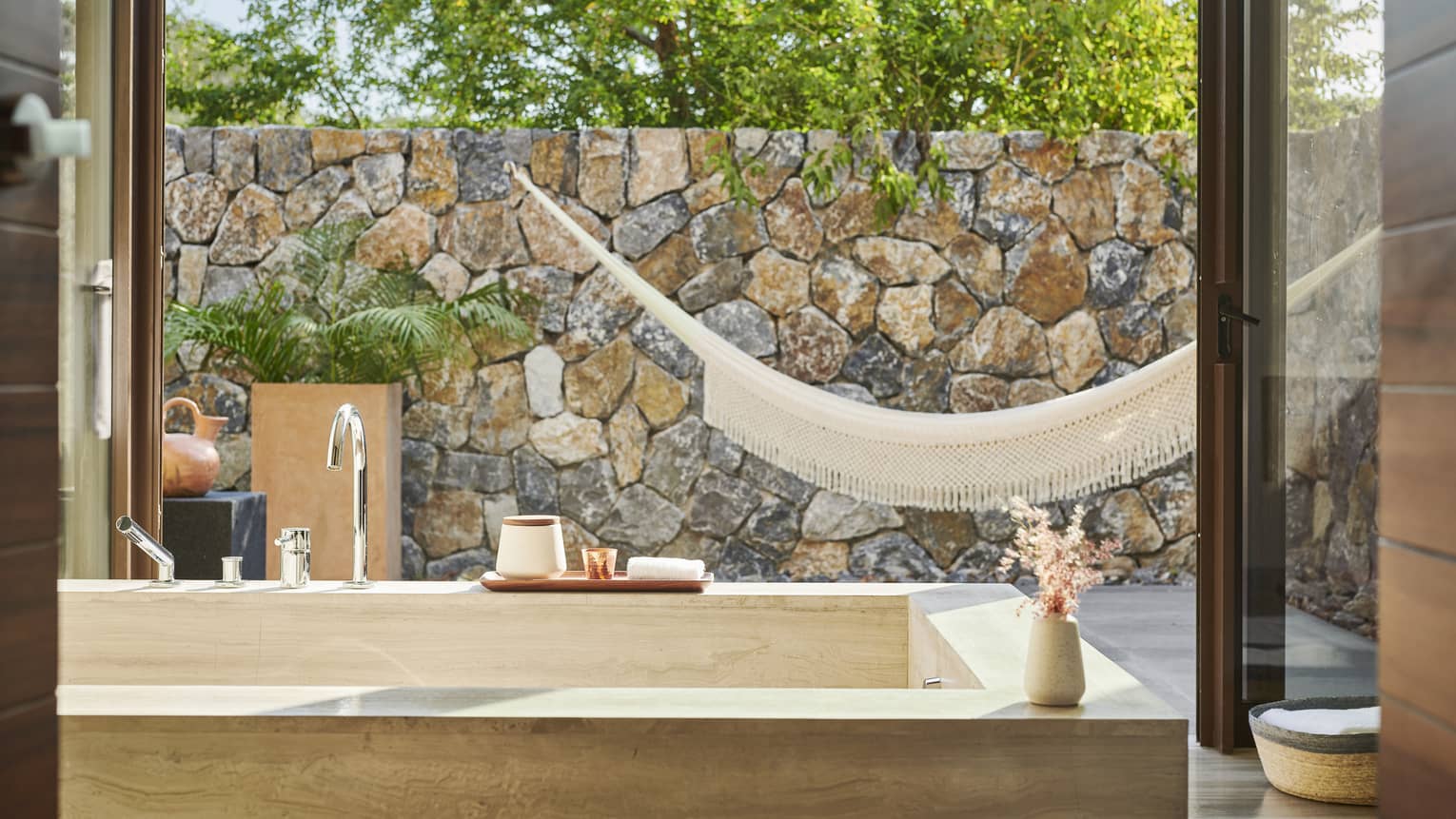 A bathroom with a large bath and large window looking outside at a hammock and stone wall.