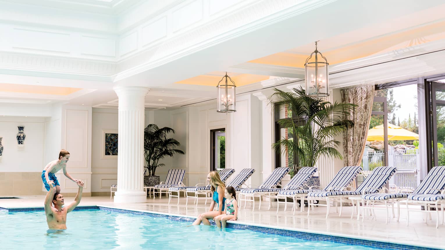 Family swimming in indoor pool, mom and daughter on ledge, boy on dad's shoulders