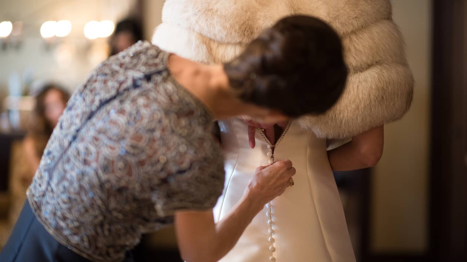 Woman helps button back of bride's wedding gown under fur coat