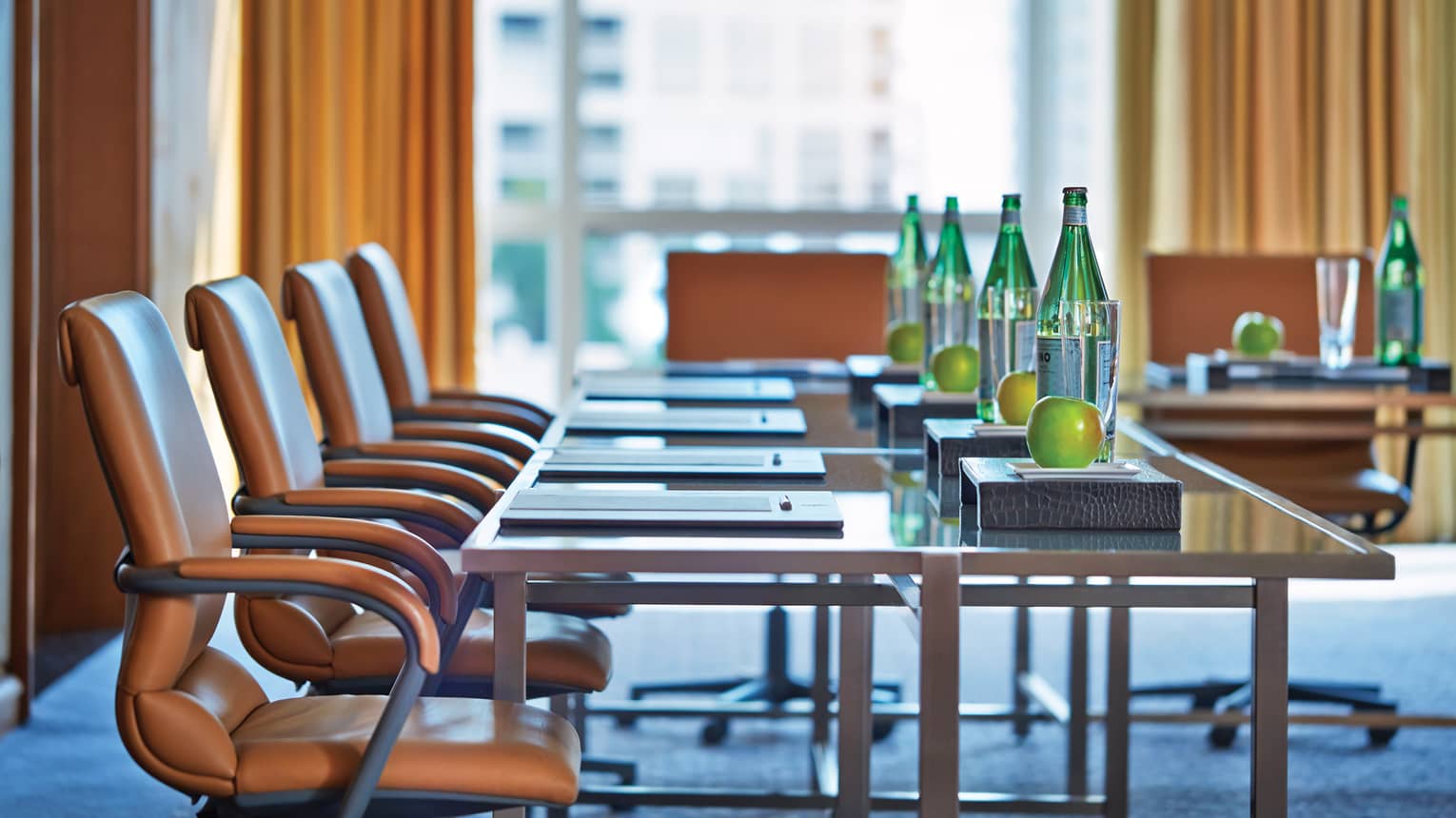 Tan leather swivel chairs line meeting table with glass bottles, apples