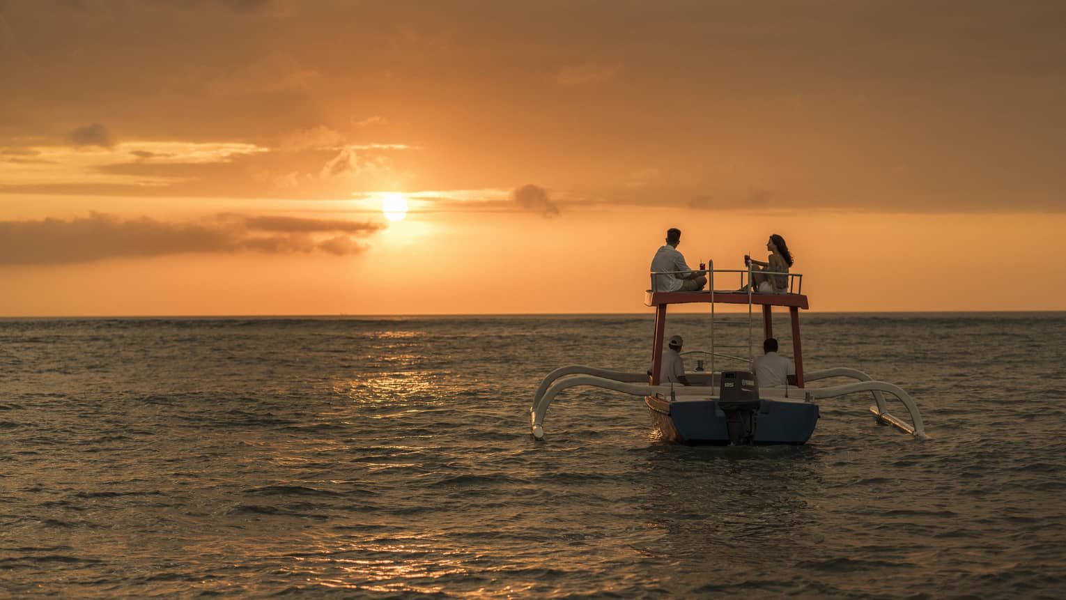 Couple sits on roof of boat sailing on ocean at sunset