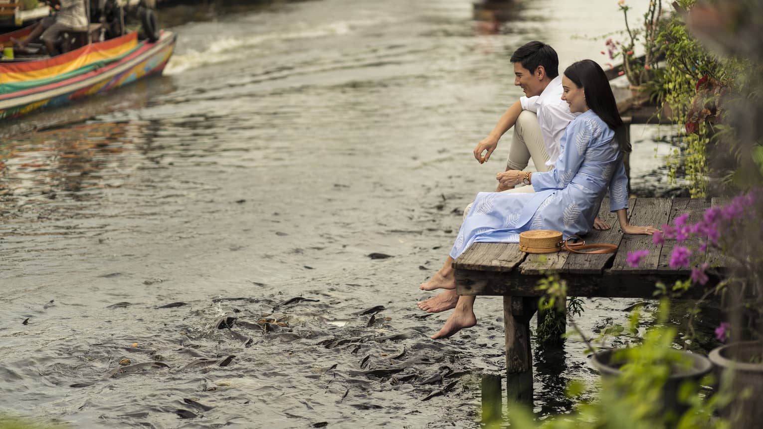Couple sits on the dock of the Chao Phraya River, hungry fish swarming beneath their dangling feet