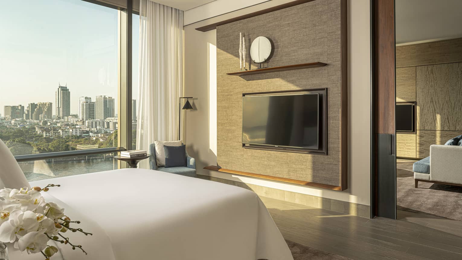 White king bed facing dark tan wall with TV, display shelf beside tall window with river and city view