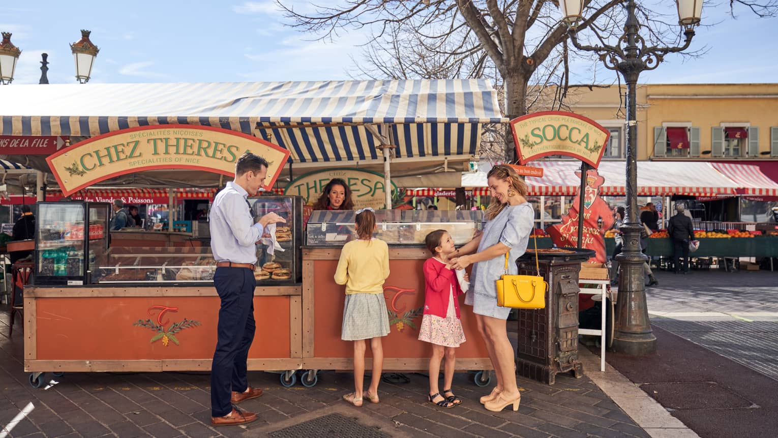 Man in blue shirt, black pants with two girls and woman in dresses at market farm stand)