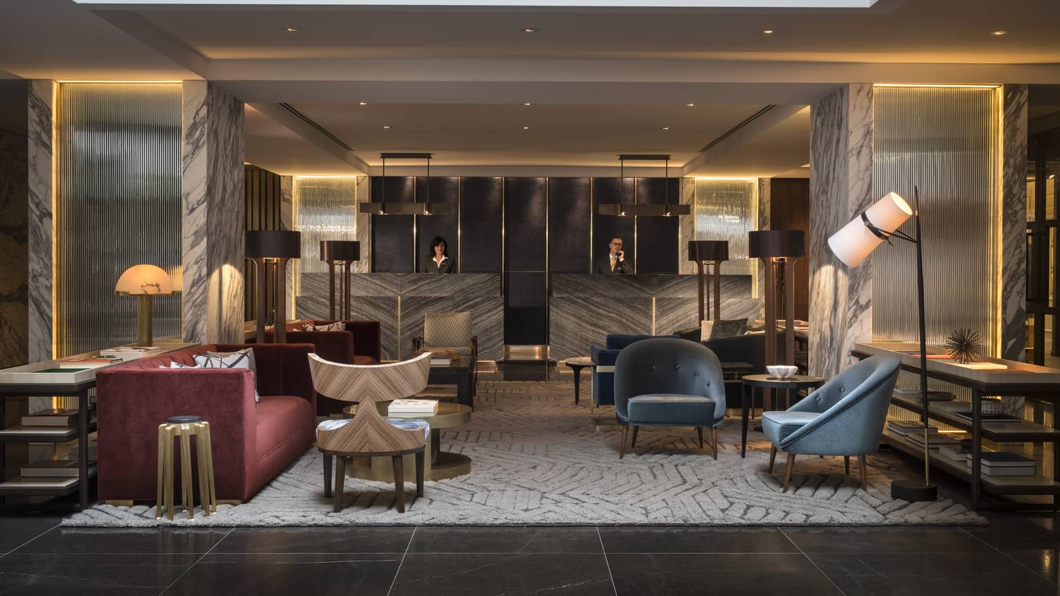 The Lobby of Four Seasons Houston is furnished with two blue velvet arm chairs, a gray and cream abstract rug, a red velvet couch, and an industrial black iron lamp