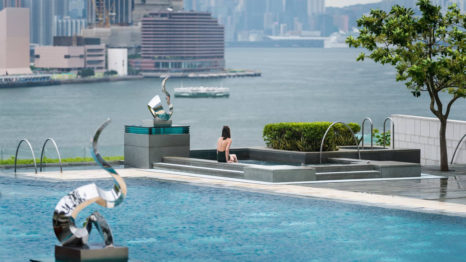 Woman in swimsuit sits on edge of swimming pool on patio near modern silver sculptures, waterfront