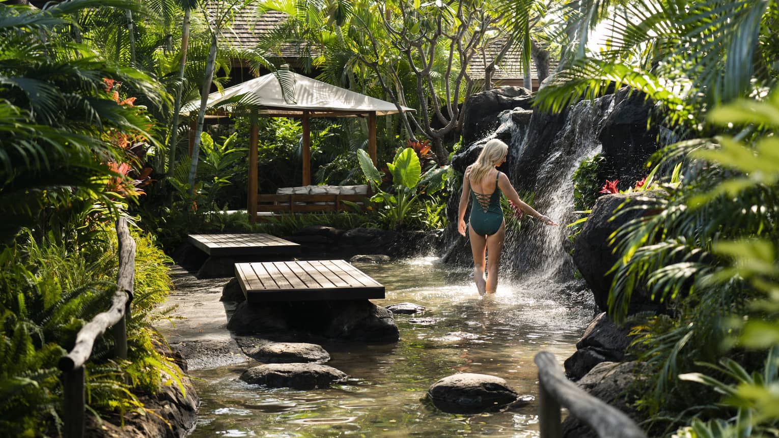 Woman wearing swimsuit stands near garden waterfall and wood platform