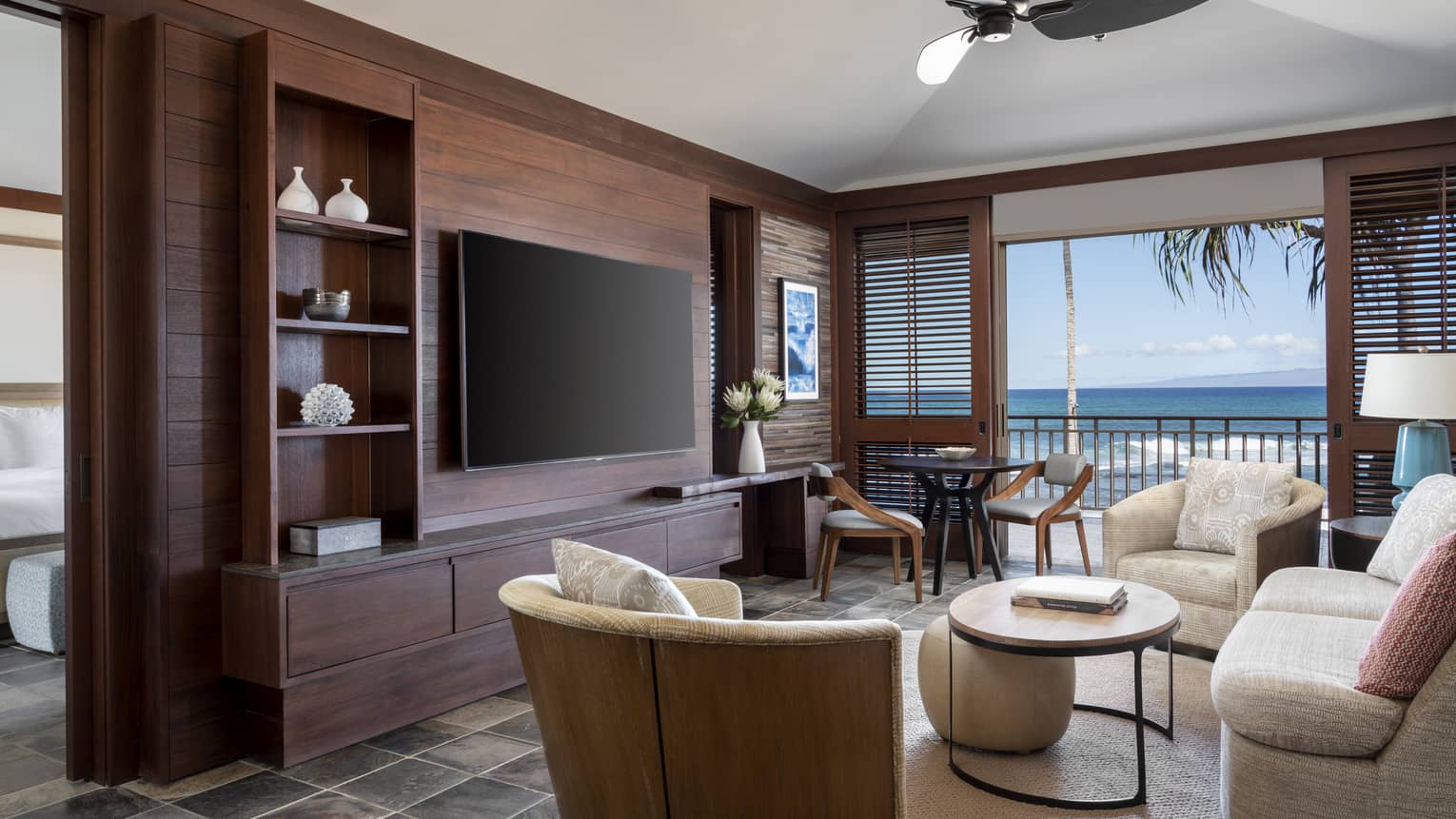 A suite's living room, with off-white sofa, two arm chairs, table, hanging flat-screen TV, walk-out balcony