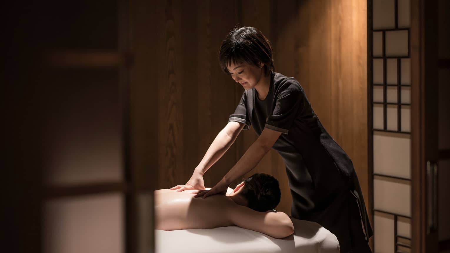 Man lies face-down on massage table as masseuse in black uniform rubs his shoulders