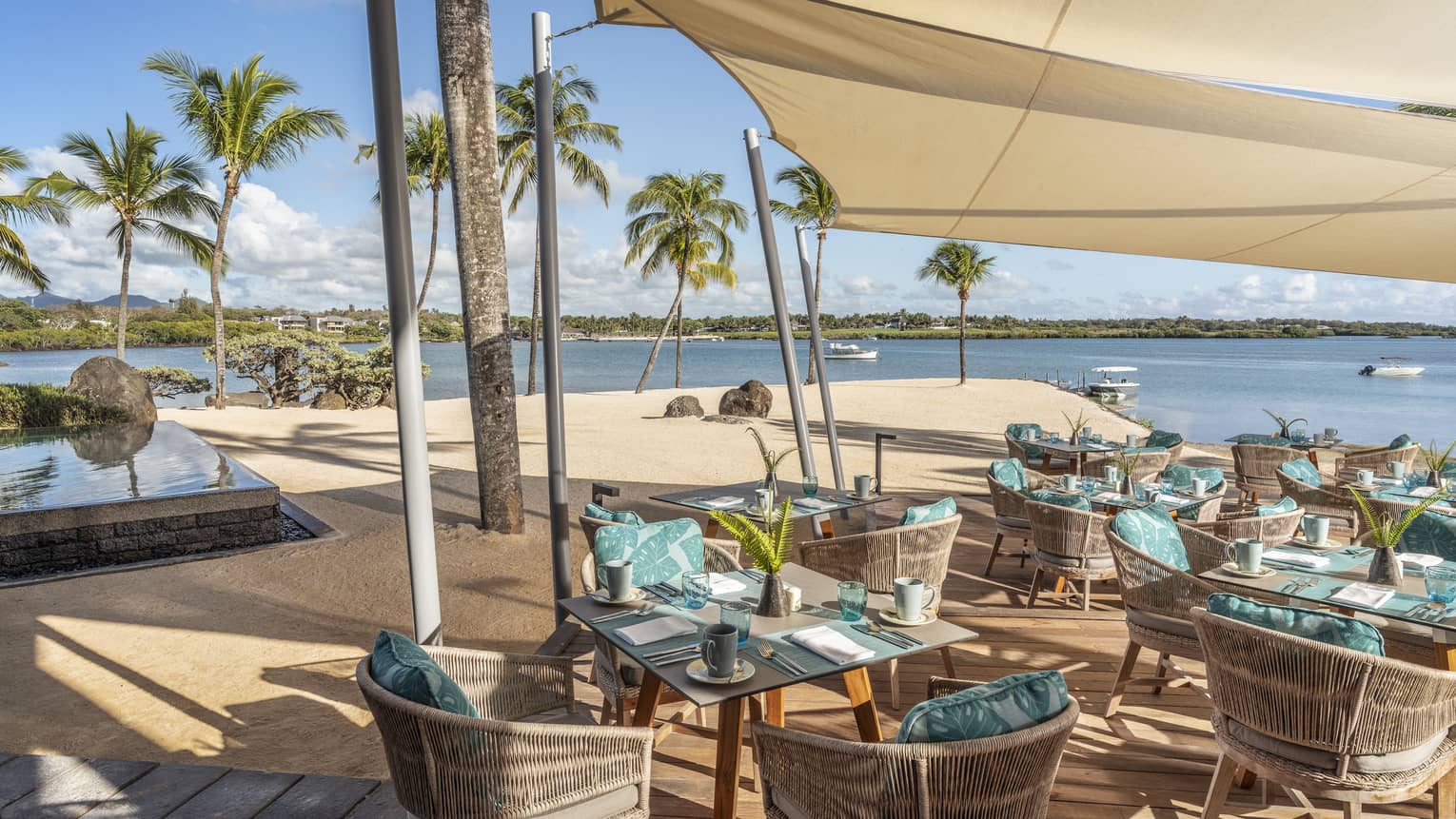 Wicker table and chair seating with aqua pillows under tan shade at waterfront