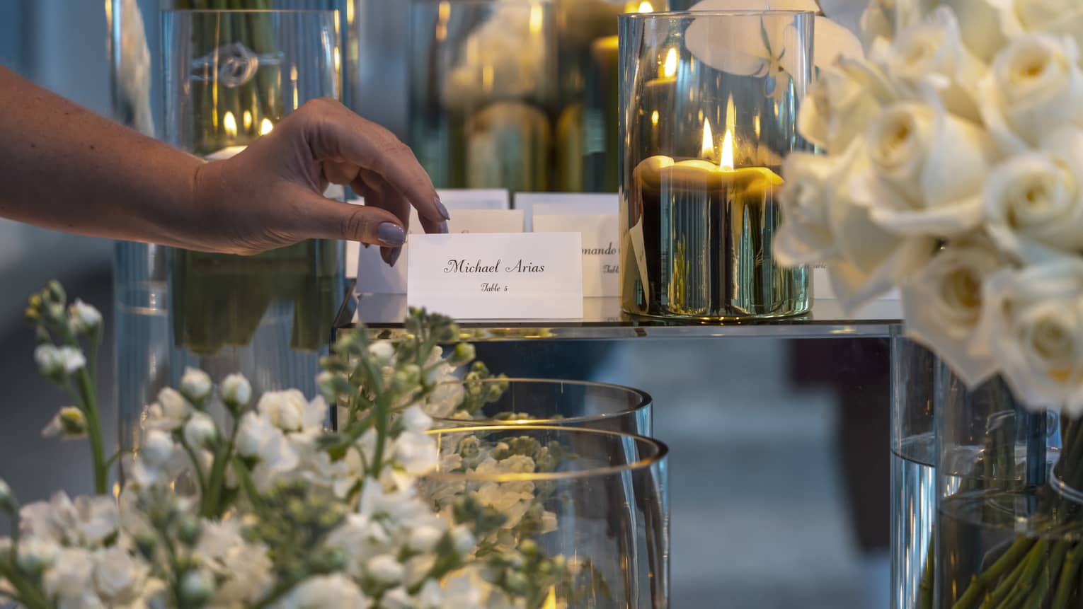 A close-up of a wedding guest picking up a place card from an elegant display at Four Seasons. The table features a sophisticated arrangement of white roses, with golden candle votives softly illuminating the background, creating a warm and welcoming ambiance for the celebration.