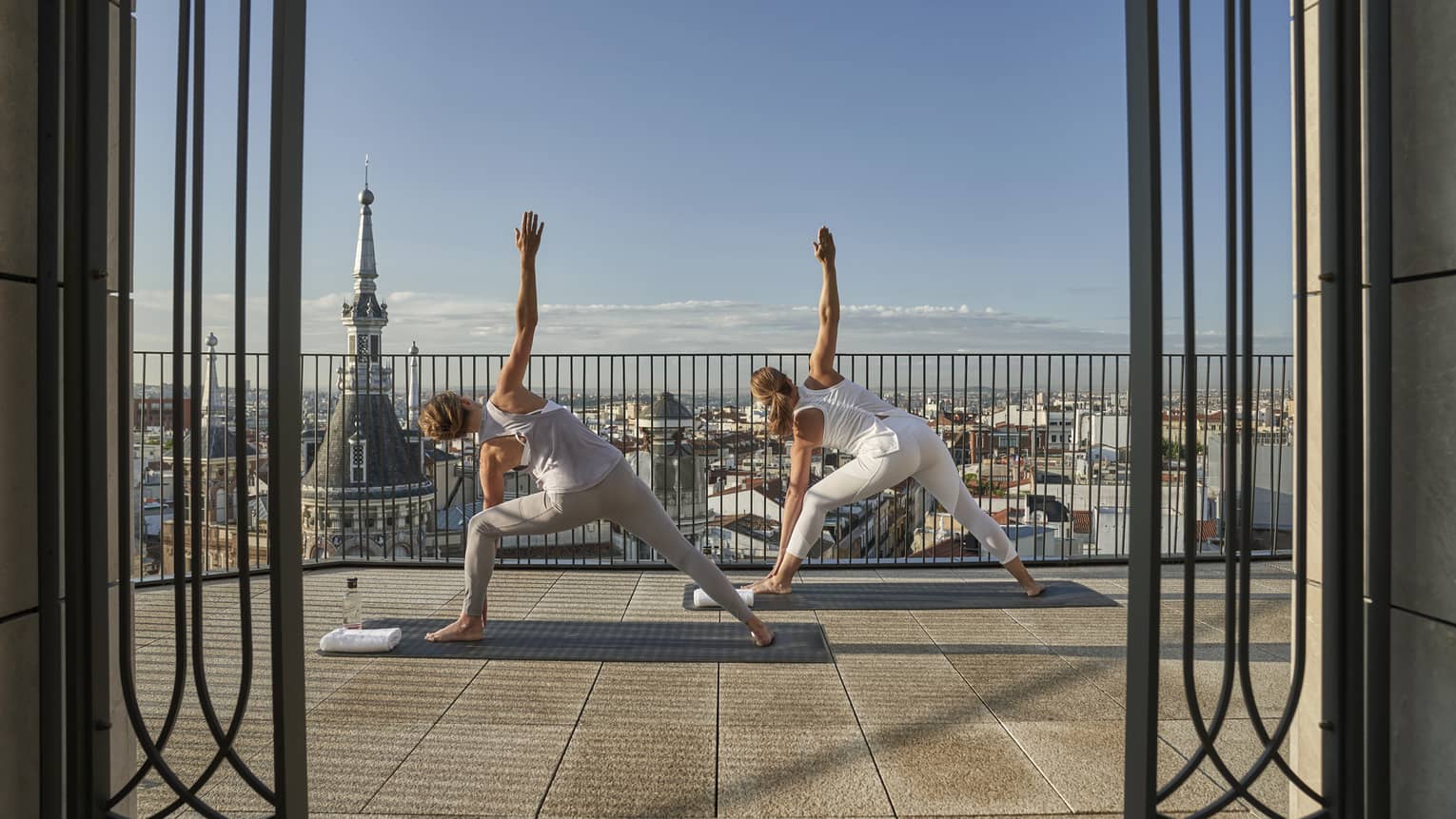 Two woman do triangle pose on outdoor terrace overlooking Madrid