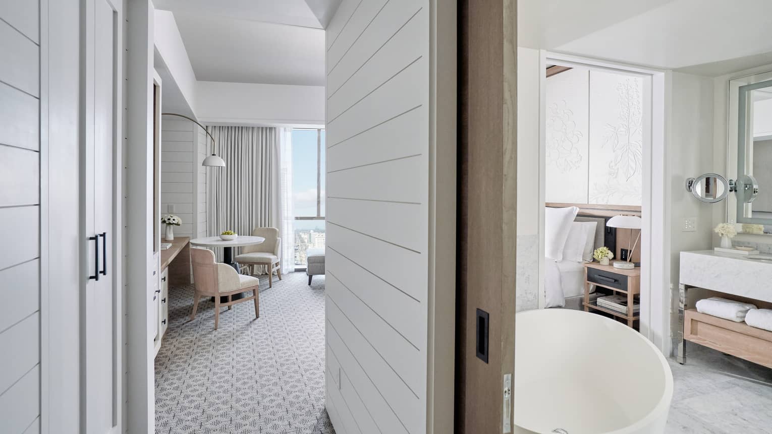 Doorway of bathroom with large soaking tub and marble countertops to the right, living area with floor-to-ceiling windows and round white table and two chairs to the left