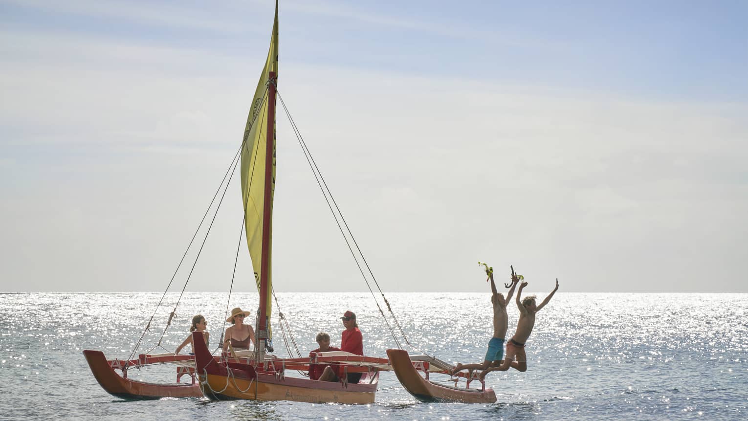 A family floats on an outrigger canoe as two people jump off the boat into the water