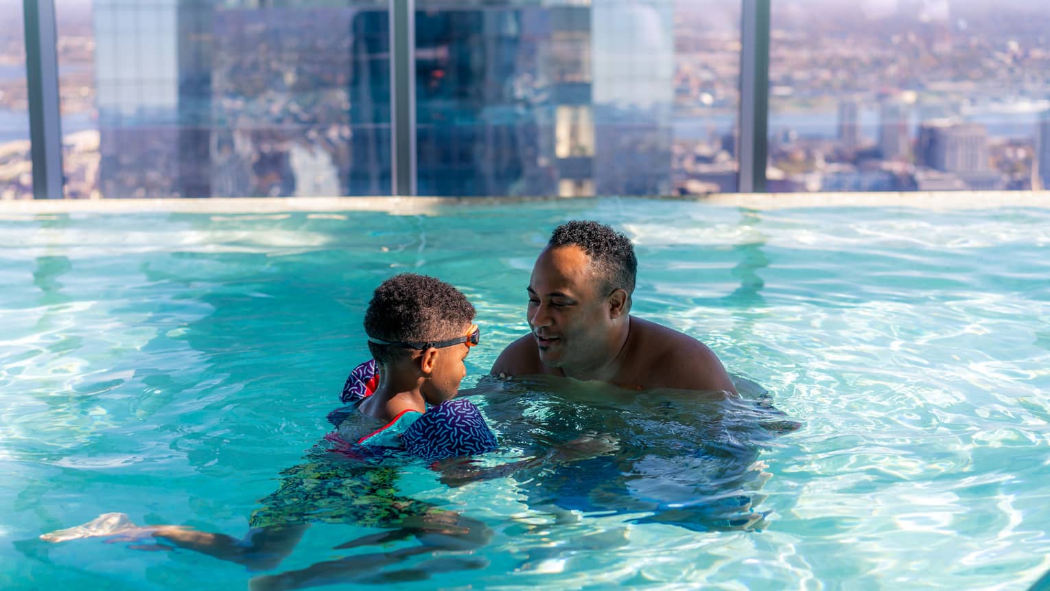 A father and son swimming in an indoor pool with large windows.
