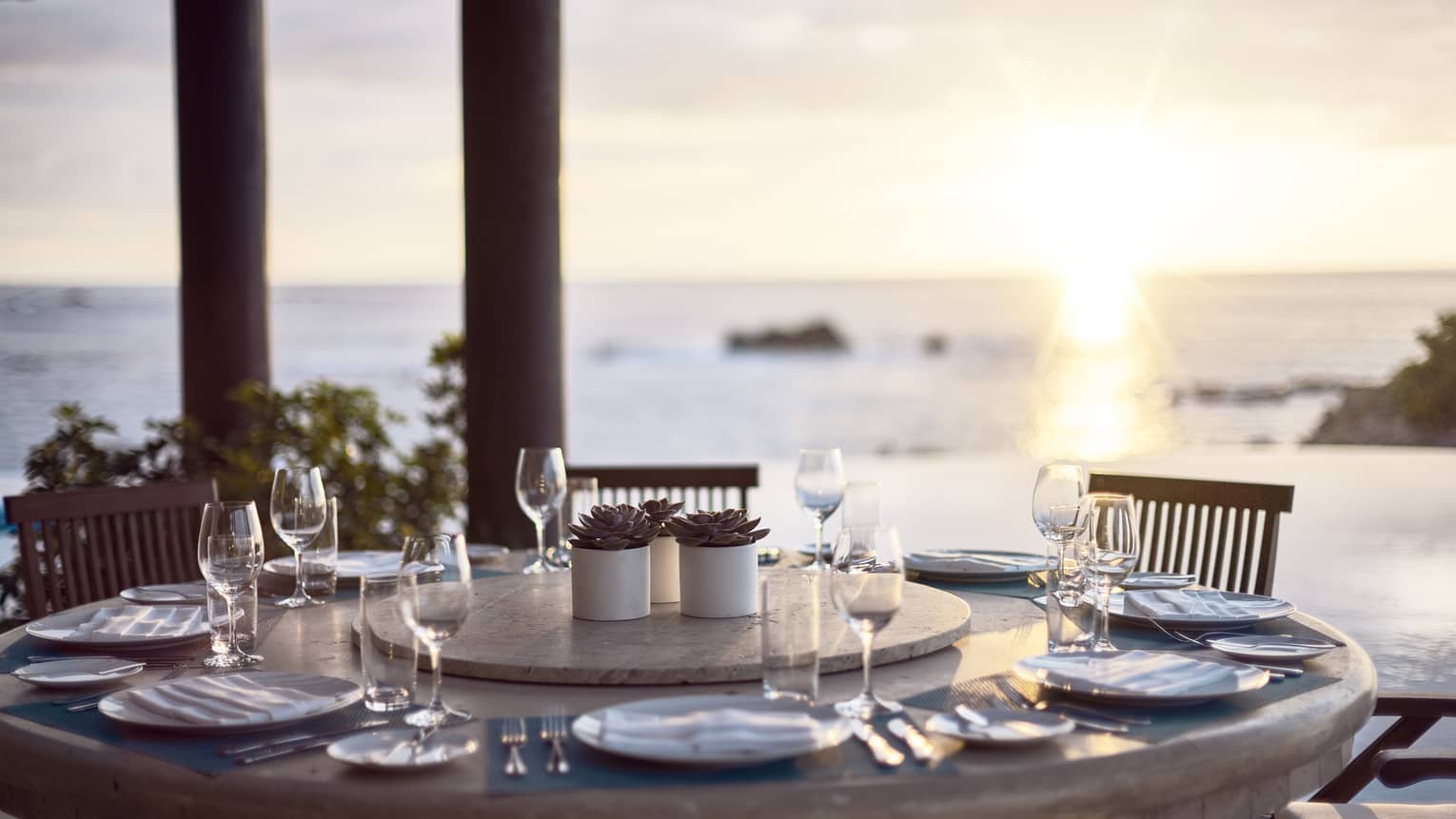 Round outdoor dining table with wine glasses, ocean and sunset in background