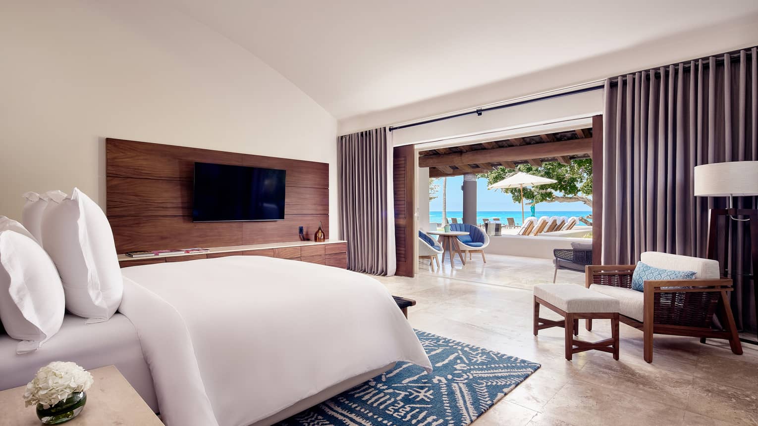 Arena Beach House bedroom with bed and white linens, white armchair, wall-mounted TV, doors open to living room