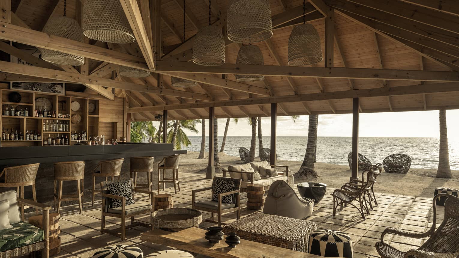 Bar with bar stools and lounge with grouped seating under vaulted wooden ceiling with open-air view of beach and ocean