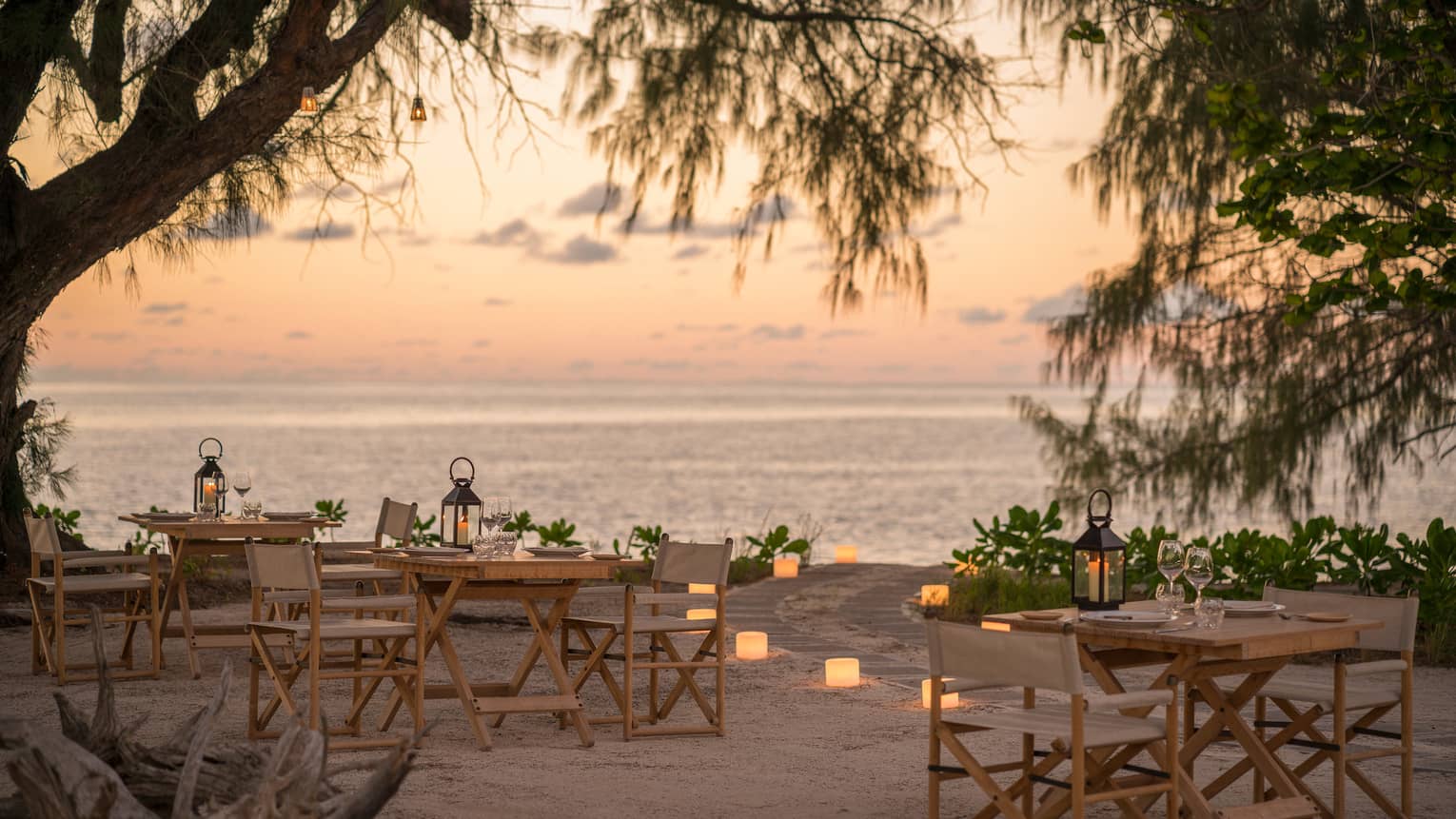 Outdoor wooden tables and chairs on sand at dusk with lit candles lining path to the ocean