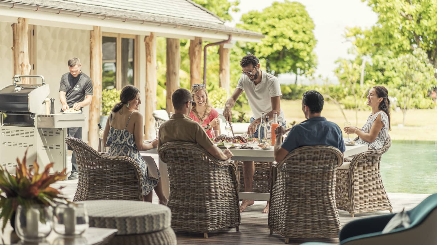 Five people seated poolside with staff member service and chef at grill