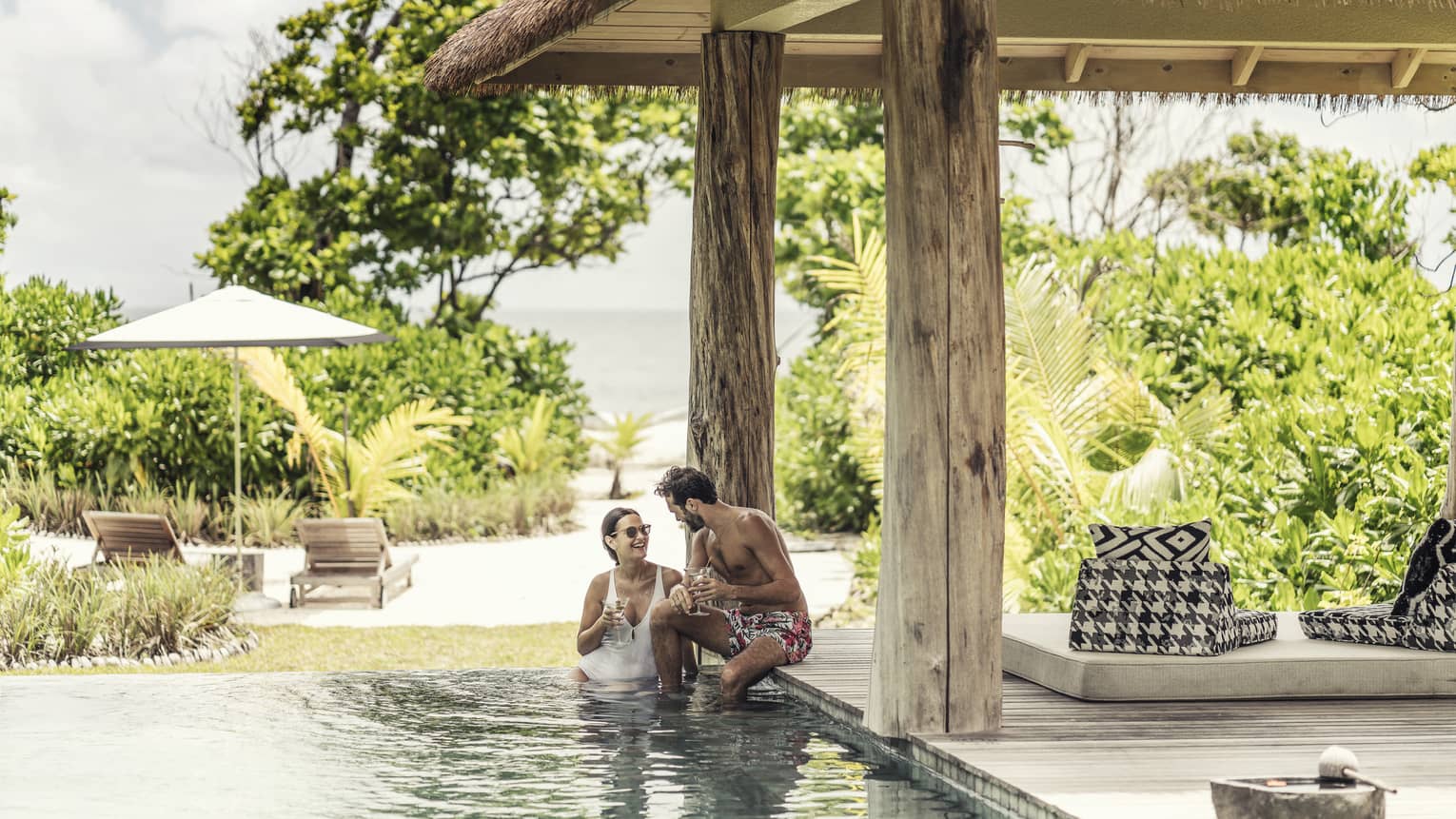 Couple in villa pool under thatched roof and view of tropical foliage