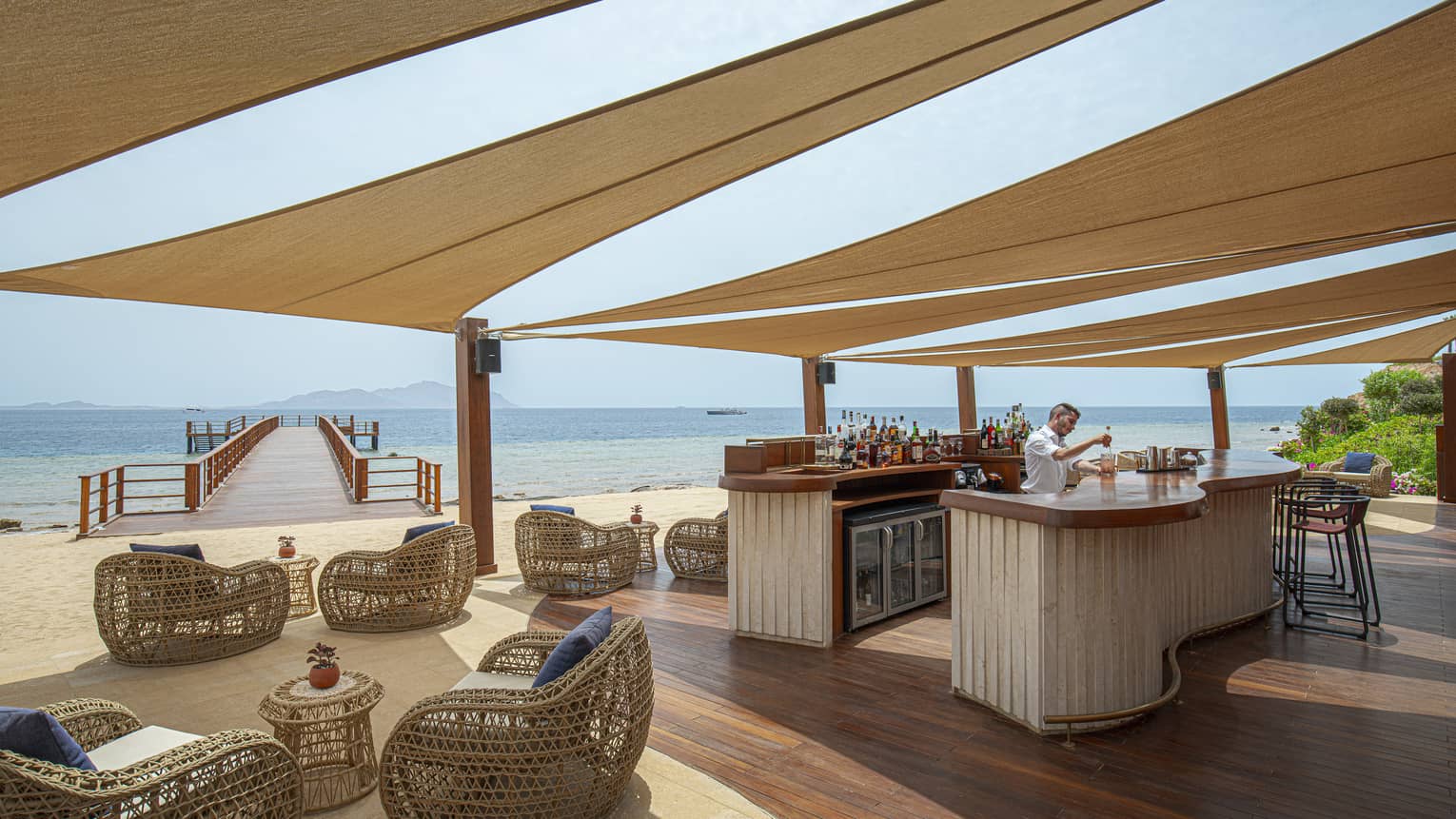 Bartender mixes drink at Breeze Bar, shaded by strips of fabric, rattan arm chairs, view of the Red Sea