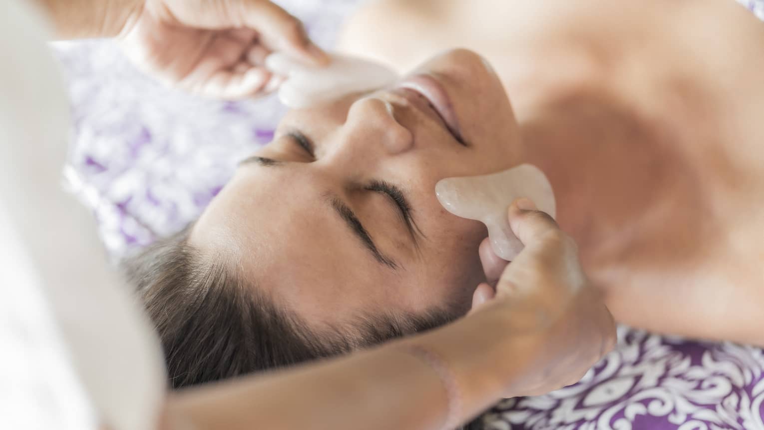 Hands hold stones at woman's cheeks on spa table