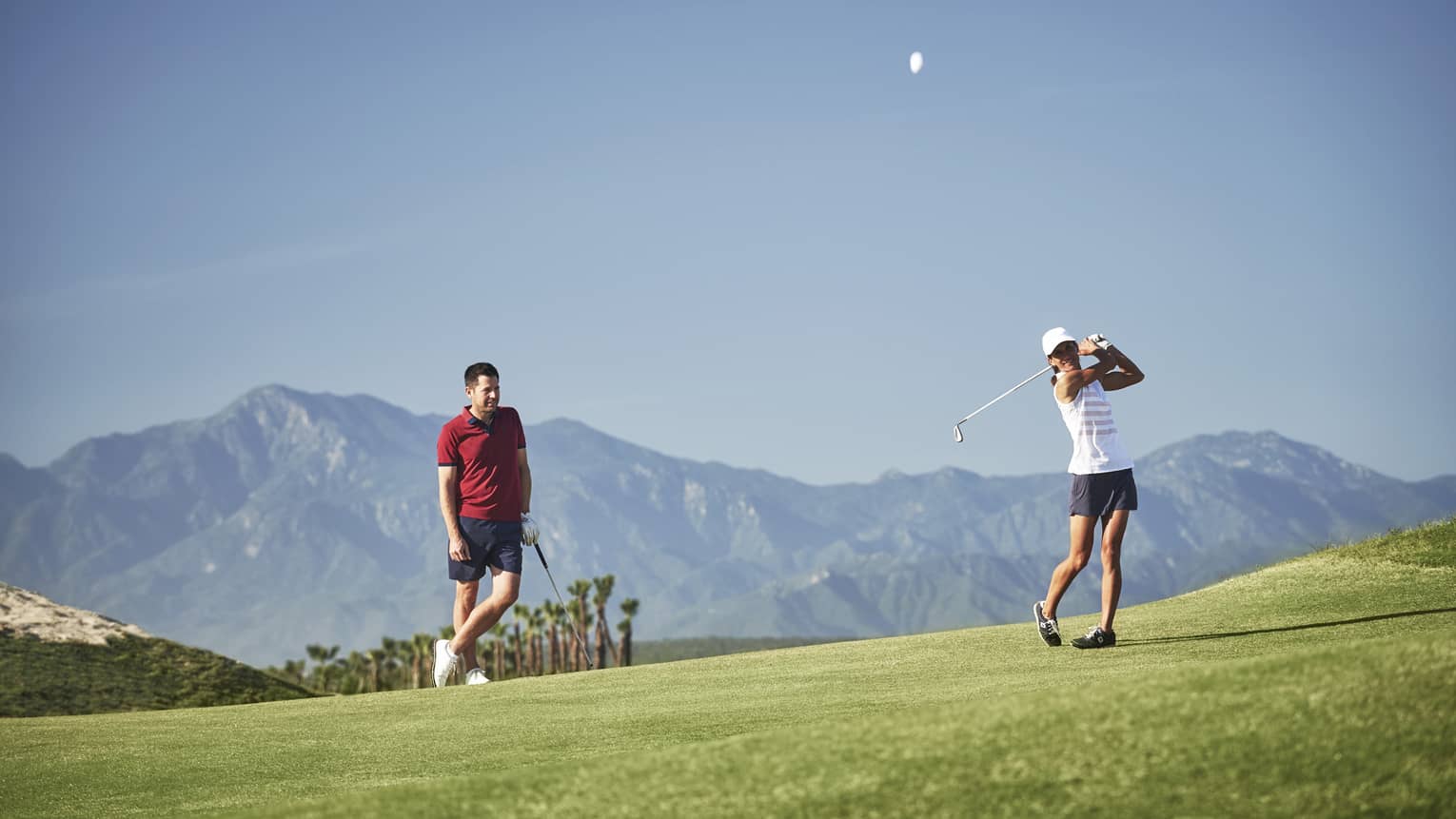 A golfer leans on their club observing another's swing on a picturesque golf course silhouetted by a mountain range.