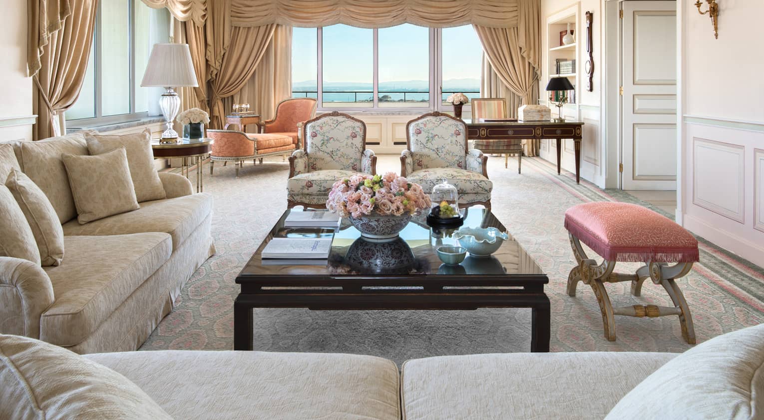 Side view of Presidential Suite seating area, coffee table, bench, two wing chairs, orange chaise, tall windows with views