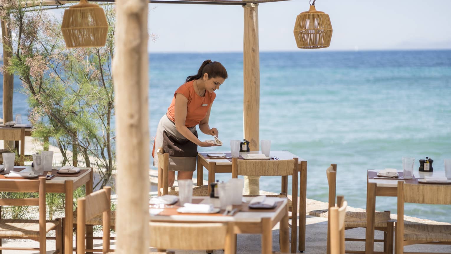 Woman in orange top sets wooden table at outdoor Taverna with wooden lanterns by ocean