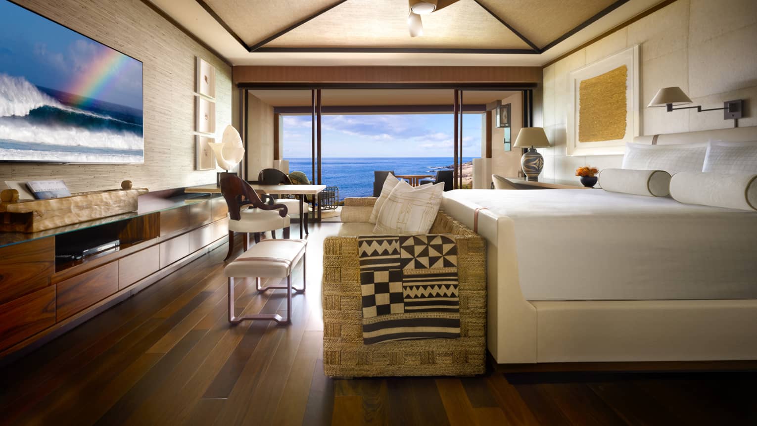 Oceanfront Room bed, small sofa and bench at foot, TV screen displaying ocean waves, rainbow