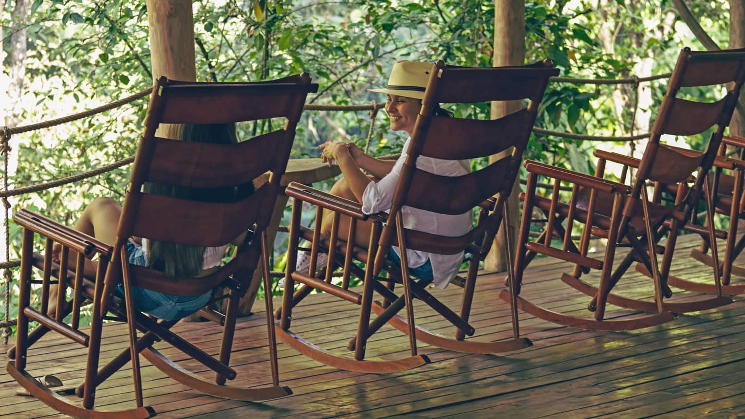 Two people sitting in large wooden rocking chairs set in a row on a wooden deck overlooking the jungle