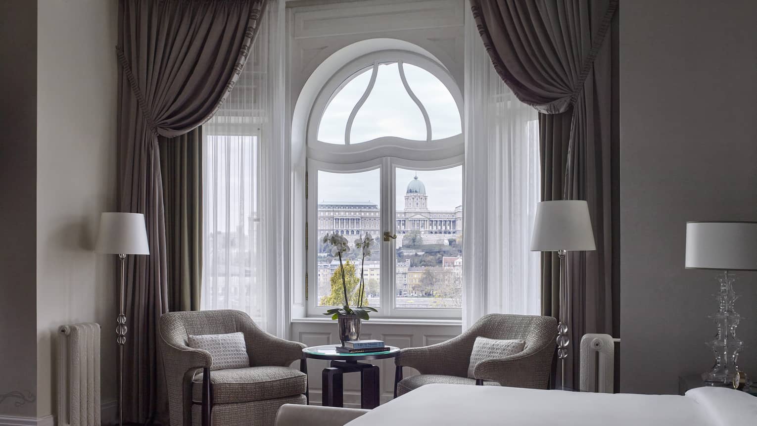 Buda Castle Presidential Suite is furnished with a two sitting chairs next to a curved window, lamps, a large bed and gray curtains. 