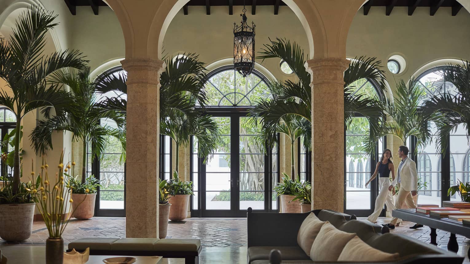 Laughing man and woman stroll through hotel lobby, decorated with modern furniture, bay windows and palm trees 