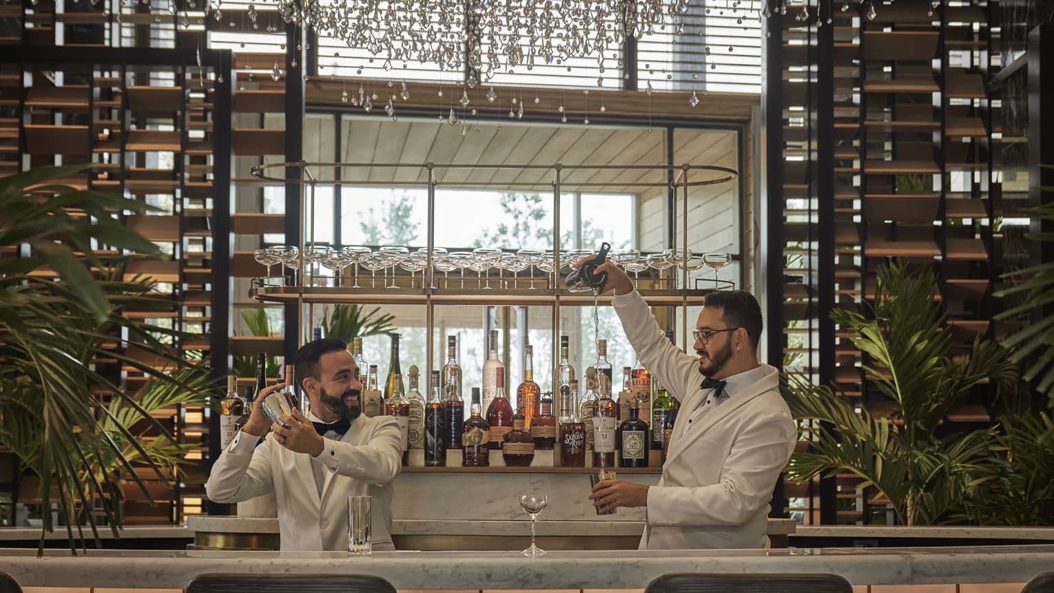 Two male bartenders clad in white prepare cocktails, liquor bottles on the bar behind them, the chandelier just visible at the top of the frame