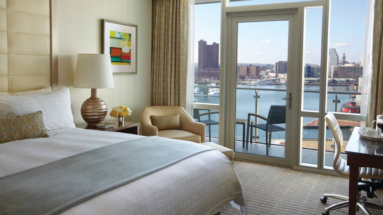 Edge of bed with folded blue blanket, armchair by glass door, balcony door, harbour view