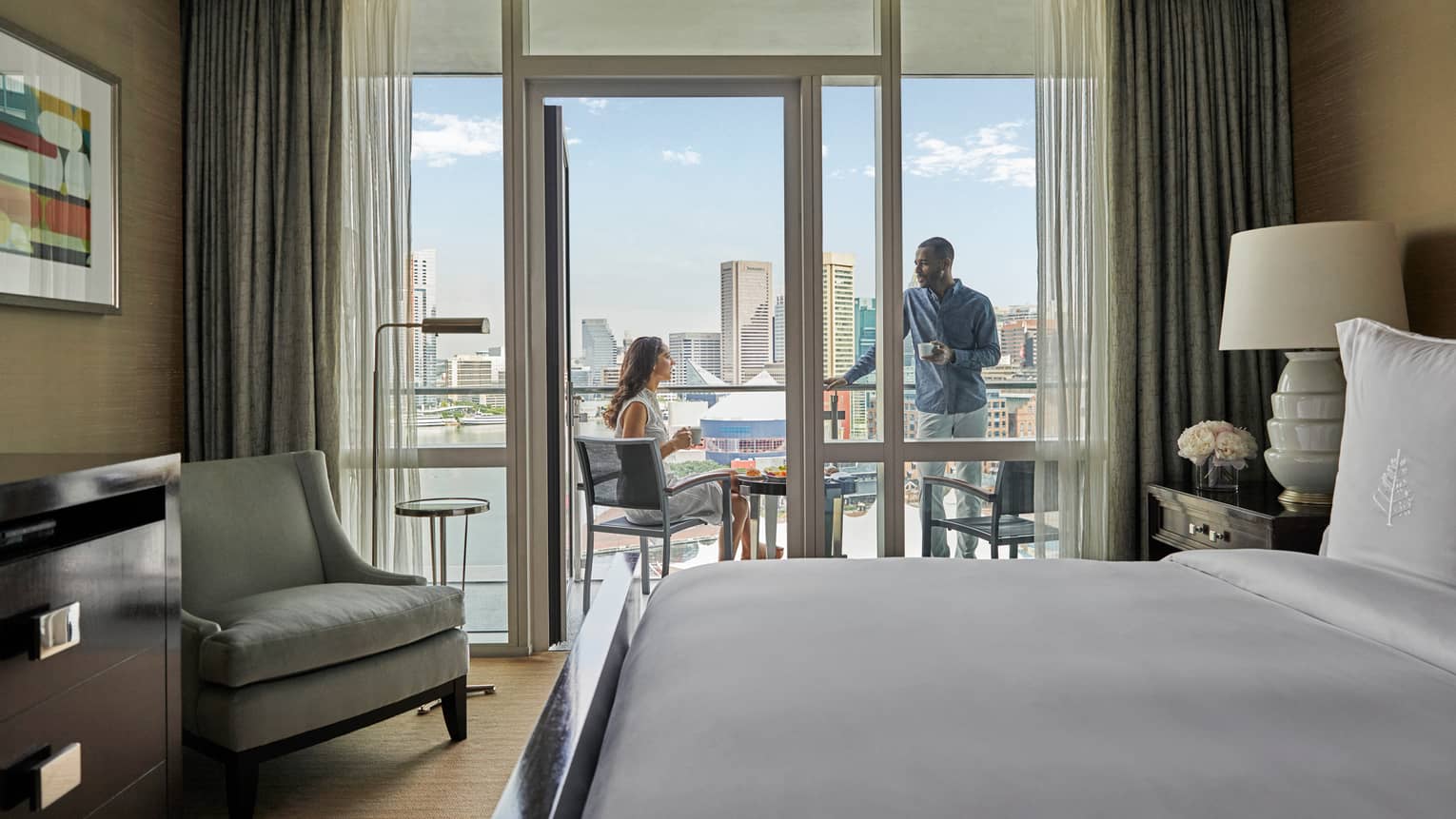 Hotel room with king bed, light grey arm chair, opening to outdoor balcony with two people talking