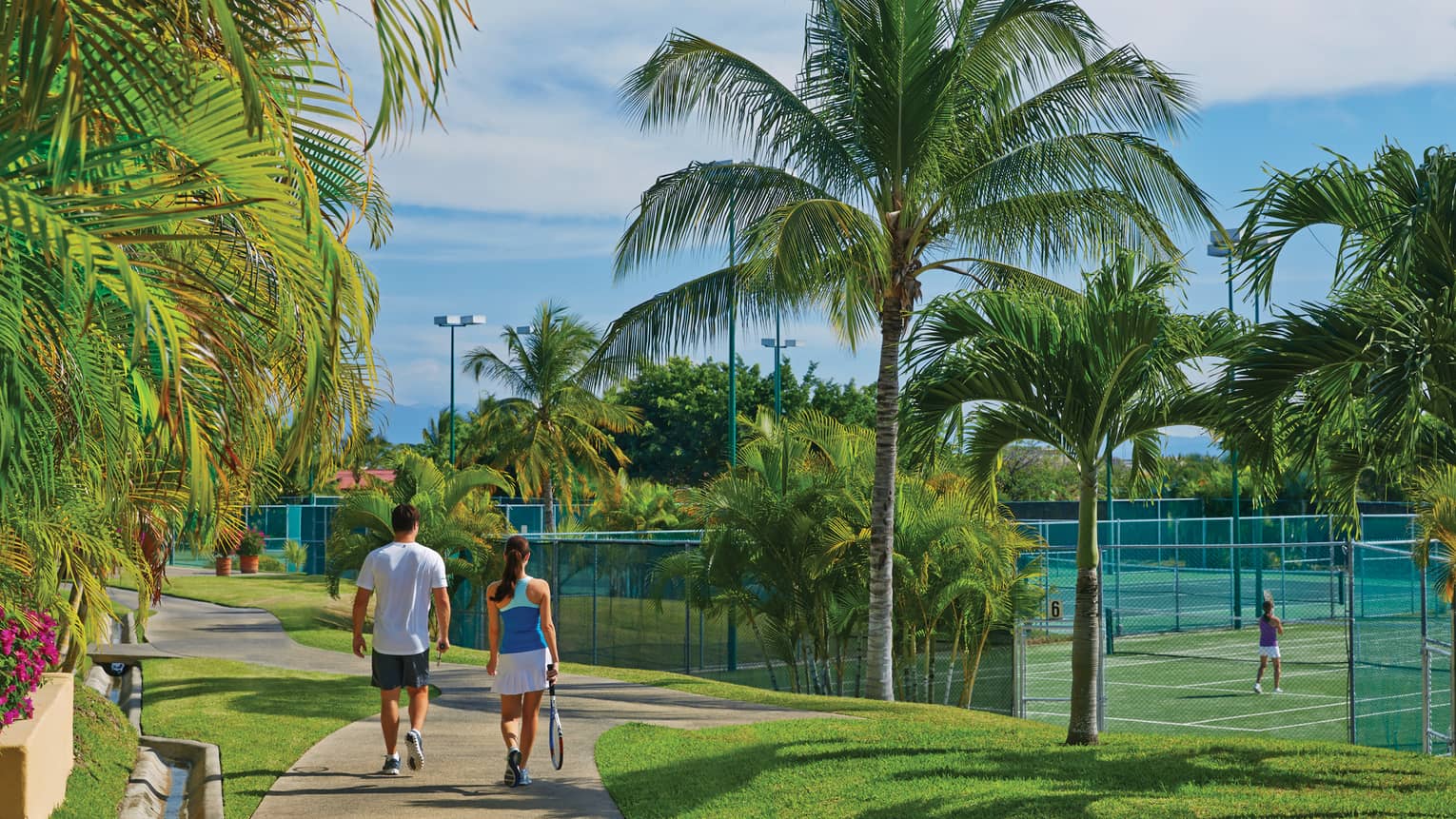 A man and woman walking along a path next to tennis courts and palm trees.