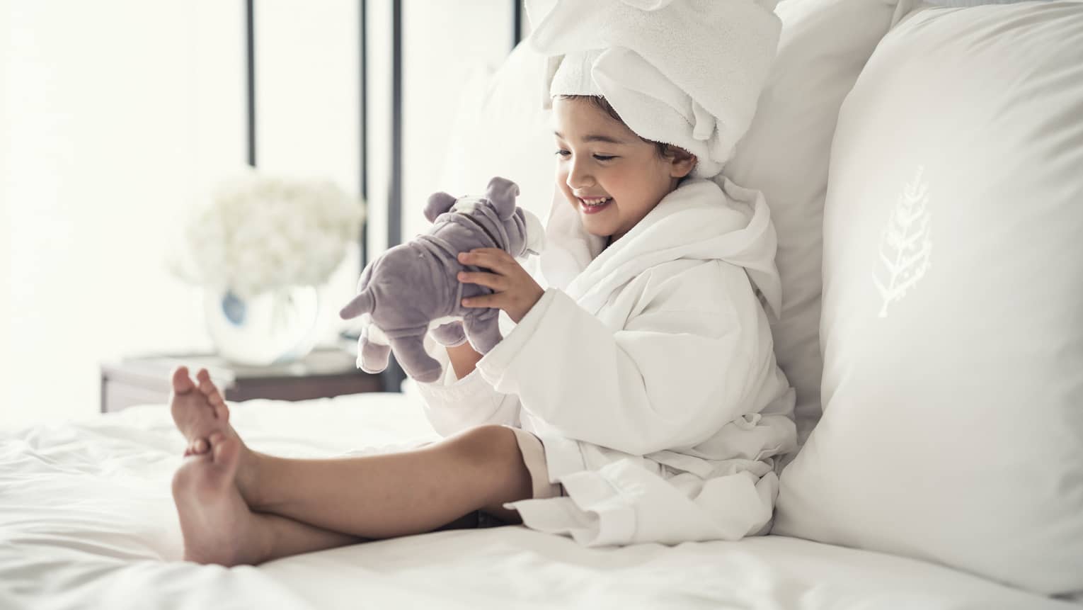 Young girl wearing a white bathrobe with towel wrap plays with plush dog toy on bed