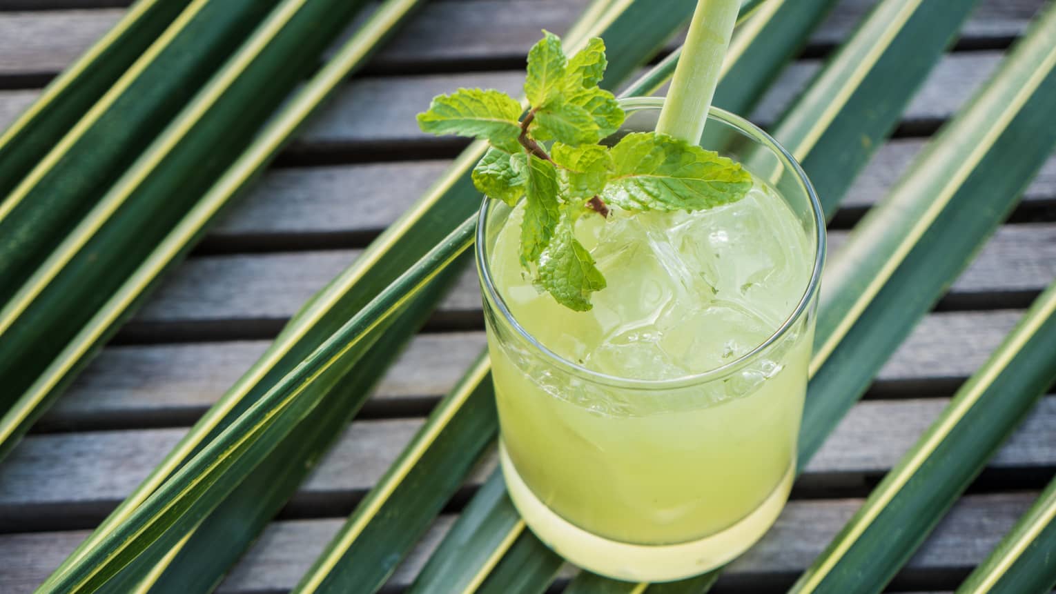 A yellow cocktail with a sprig on mint in it resting on greenery on a table