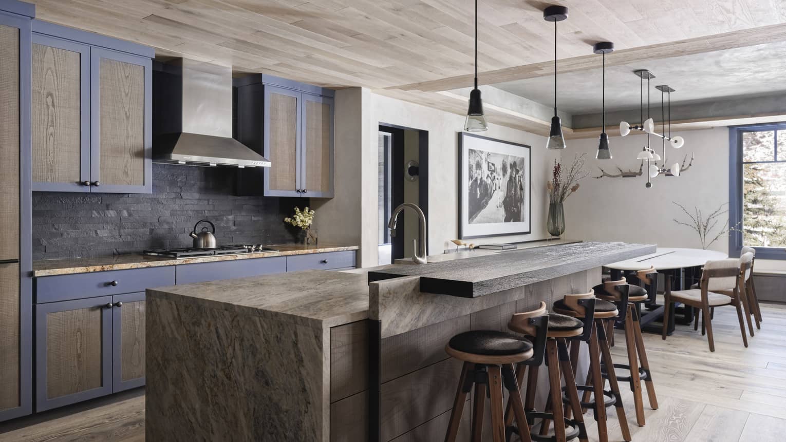 Residence kitchen with grey wood cabinetry, long centre island with leather barstools, stovetop range, dining table on right