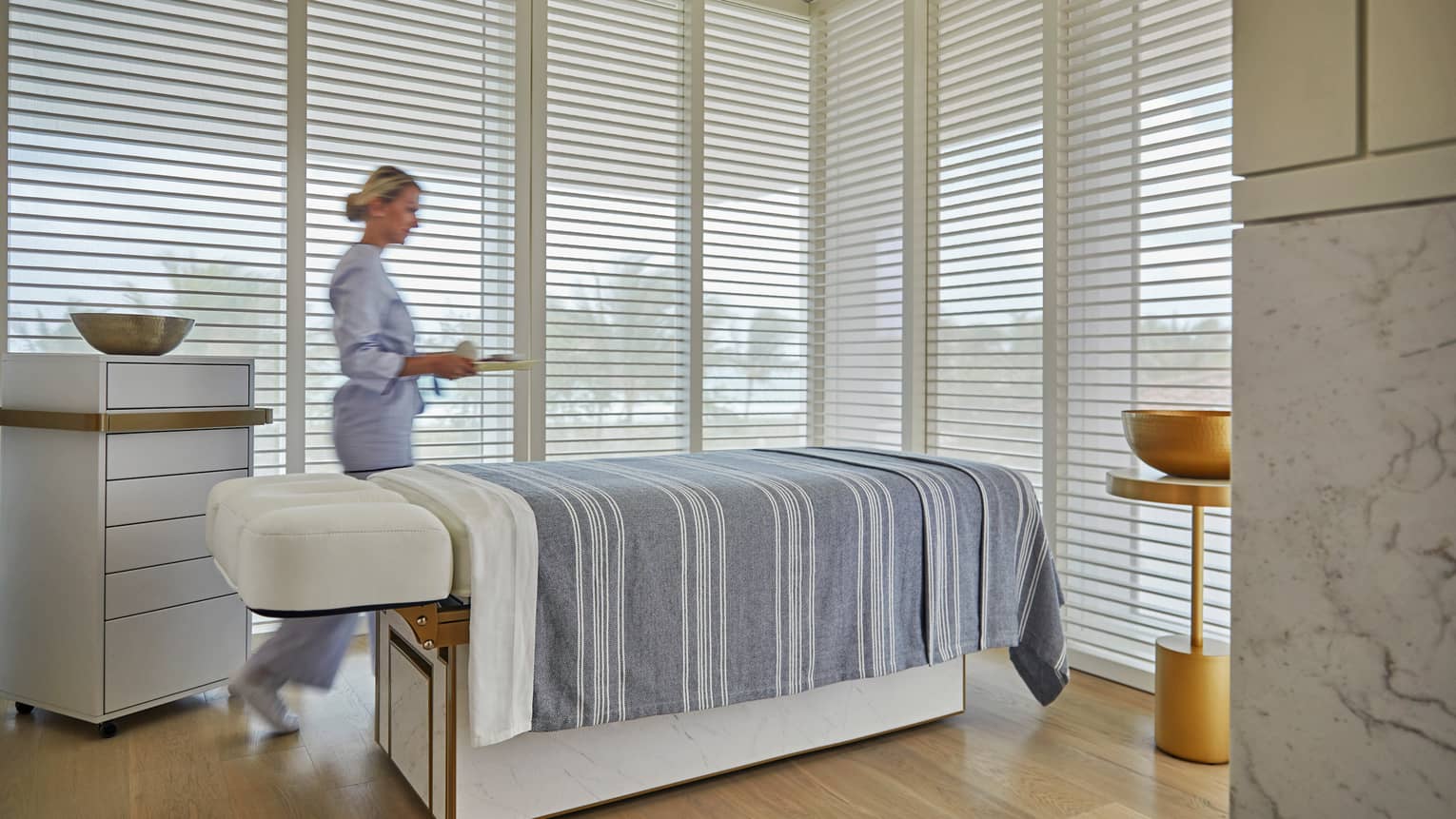Spa attendant carries tray in treatment room, wall-to-ceiling windows, massage table covered with striped sheet 