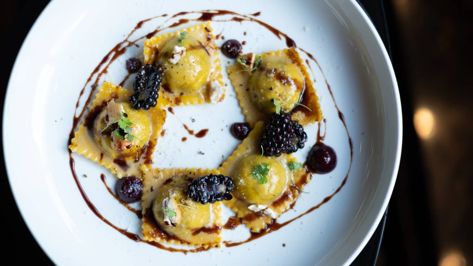 Five ravioli pastas arranged in circle on dish, garnished with sauce and blackberries