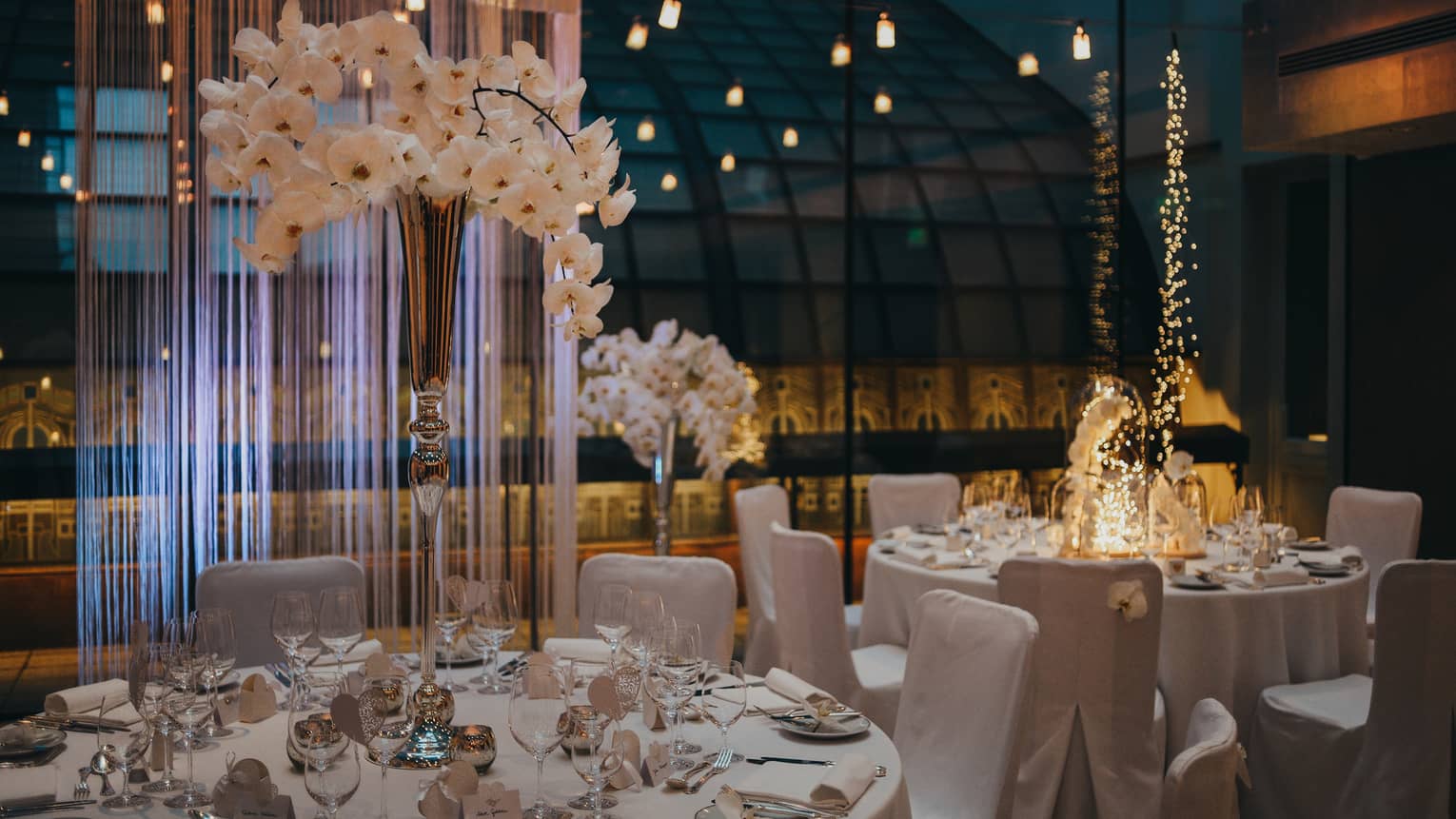 White dining tables and chairs with vase of orchids beside glass wall and reflecting lights