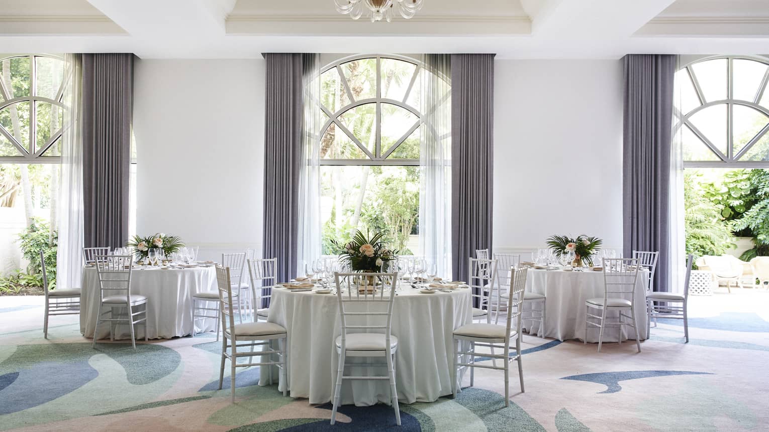 A large room with colourful carpet, round tables with white cloths, and large windows.