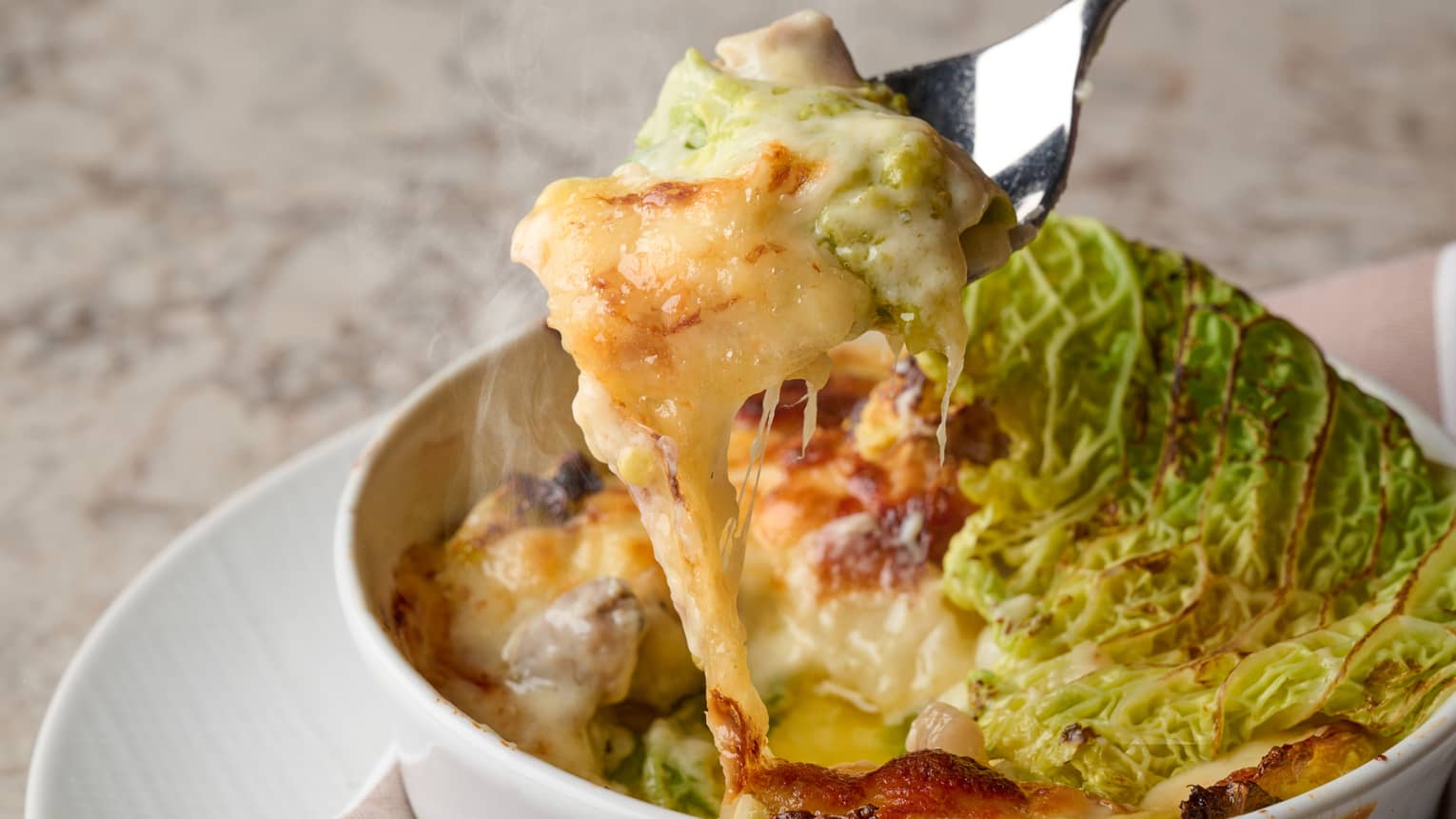 A steaming forkful is pulled from a ramekin of rich, cheesy casserole garnished with a charred green Savoy cabbage leaf.