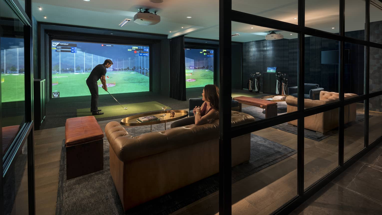 A man uses an indoor golf simulator while a woman watches from a nearby couch