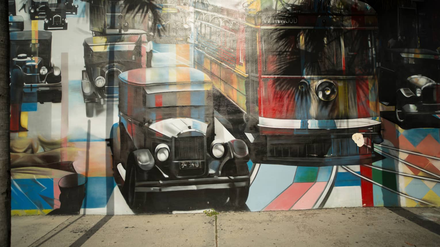 Palm trees cast shadows on a mural of vintage cars and a bus painted in a kaleiscopic pattern of colours and grey shading.
